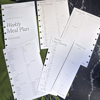 Jane’s Agenda Weekly Meal Plan planner inserts arranged in an overlapping flat lay on a black marble surface with green fabric accents. The collection features a Weekly Meal Plan title page, Monday-through-Sunday meal planning layouts with spaces for breakfast, lunch, dinner, and snacks, categorized grocery lists, dot-grid notes sections, and preprinted pantry staples checklists. The minimalist black-and-white inserts feature discbound punch holes and are designed to simplify weekly meal planning, grocery s