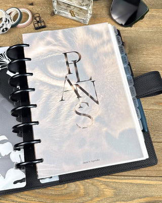 Lifestyle photo of a discbound planner opened to a translucent vellum dashboard featuring the word “PLANS” over a softly faded tiger face. The planner sits in a black cover with monthly tabs visible, surrounded by stationery accessories on a wooden desk.