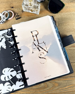Lifestyle photo of a discbound planner featuring a tropical dashboard with layered monstera and banana leaves surrounding a misty palm forest. Styled on a wooden desk with black discs, a blue pen, sunglasses, washi tape, and desk accessories for a fresh, nature-inspired planning setup.