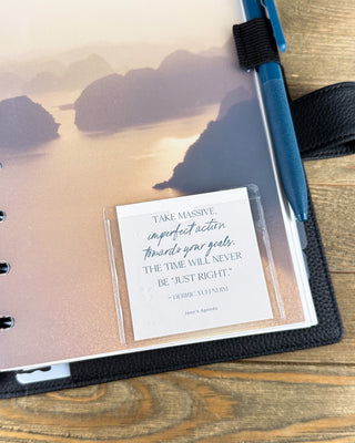 Clear adhesive planner card pocket attached to a discbound planner dashboard, displaying an inspirational quote card against a golden tropical landscape with a blue pen and black leather planner cover.