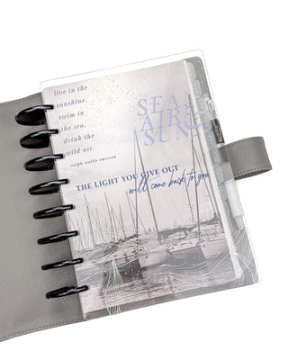 The gray leather cover with a planner featuring a marina of boats layed with the Sea, Air & Sun planner cover. 