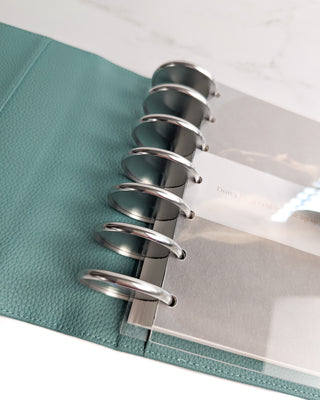 Close-up of a sea glass color leather planner cover with a discbound planner and silver metal binding discs on a white background