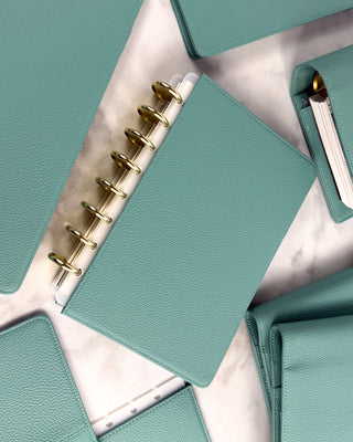 Light teal colored vegan leather discbound planner cover with Champagne metal discs,  surrounded by othe sea glass leather discbound covers.