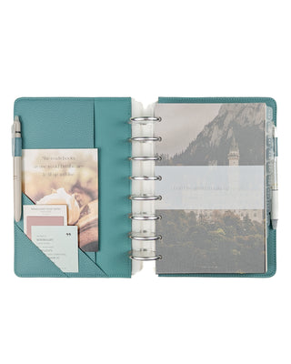 Light teal open book discbound planner with decorative accessories and silver metal binding discs over a white background