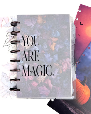 Open Jane’s Agenda discbound planner with dark metallic discs and monthly side tabs, featuring a frosted dashboard with the phrase “YOU ARE MAGIC.” layered over a colorful autumn leaf design, with coordinating Halloween dashboards beside it.