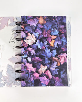 Open Jane’s Agenda discbound planner with dark metallic discs and monthly side tabs, featuring a vibrant dashboard covered in layered autumn leaves in deep purple, blue, pink, and burgundy tones.