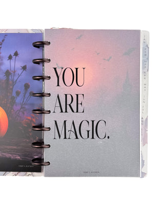 Open Jane’s Agenda discbound planner with dark metallic discs and monthly side tabs, featuring a frosted dashboard with the phrase “YOU ARE MAGIC.” layered over a moody pink and purple Halloween scene with flying bats.