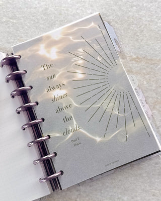 Open Jane’s Agenda discbound planner with metallic purple discs and monthly side tabs, featuring a frosted dashboard with a sunburst design and the quote, “The sun always shines above the clouds,” layered over a glowing gold water-pattern dashboard.