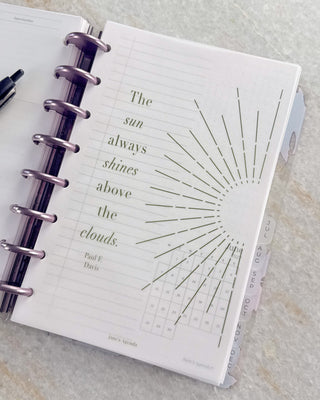 Open Jane’s Agenda discbound planner with metallic purple discs and monthly side tabs, featuring a translucent dashboard with a sunburst design and the quote, “The sun always shines above the clouds,” layered over lined and calendar planner pages.