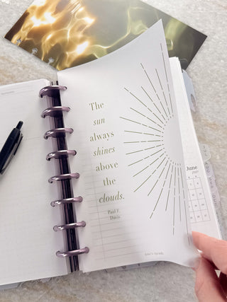 Open Jane’s Agenda discbound planner with metallic purple discs and monthly side tabs, featuring a frosted dashboard with a sunburst design and the quote, “The sun always shines above the clouds,” as a hand lifts the dashboard to reveal planner pages underneath.