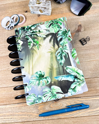 Lifestyle photo of a discbound planner featuring a lush tropical dashboard with layered monstera and banana leaves framing a misty palm forest scene. The planner is styled on a wooden desk with black discs, a blue pen, sunglasses, washi tape, and desk accessories, creating a fresh, nature-inspired planning aesthetic.