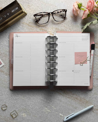 Featuring a junior-size weekly planner layout with two-inch silver binding discs pale blush pink cover, monthly dividers with white ink, and a pale pink highlighter in the penloop. Set on a marble table with flowers, glasses, a gold calendar, paperclips, and a pen for planning. 