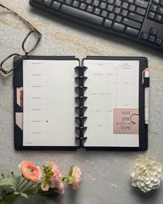Featuring a junior-size weekly planner layout with two-inch silver binding discs pale blush pink cover, monthly dividers with white ink, and a pale pink highlighter in the penloop. Set on a marble table with flowers, glasses, a gold calendar, paperclips, and a pen for planning. 