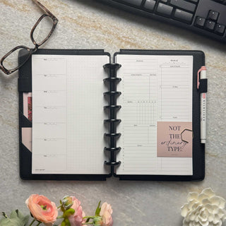Featuring a junior-size weekly planner layout with two-inch silver binding discs pale blush pink cover, monthly dividers with white ink, and a pale pink highlighter in the penloop. Set on a marble table with flowers, glasses, a gold calendar, paperclips, and a pen for planning. 