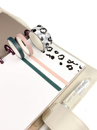 Three rolls of washi tape rest on a white planner page inside a blond leather discbound planner. The set includes one wide roll with a black leopard print design and two coordinating slim solid-color rolls, creating a stylish, safari-inspired planning accent.