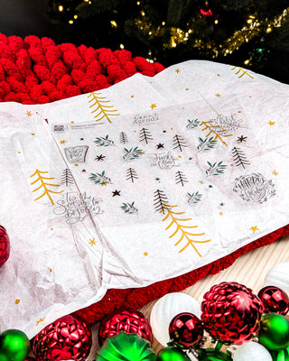 Jane’s Agenda Winter Wishes decorative sticker sheet featuring festive holiday illustrations of minimalist evergreen trees, botanical sprigs with red berries, scattered gold and black stars, and elegant hand-lettered phrases including “’Tis the Season to Be Jolly,” “Jingle All the Way,” “Let It Snow,” and “Happy Holidays.” The sticker sheet is displayed on white tissue paper with gold holiday accents, surrounded by a cozy red knitted blanket, colorful Christmas ornaments, and twinkling tree lights.