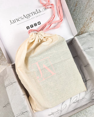 Jane’s Agenda gift packaging featuring a natural cotton drawstring pouch with a blush pink JA logo, wrapped in blush ribbon and presented inside a branded white gift box with tissue paper.