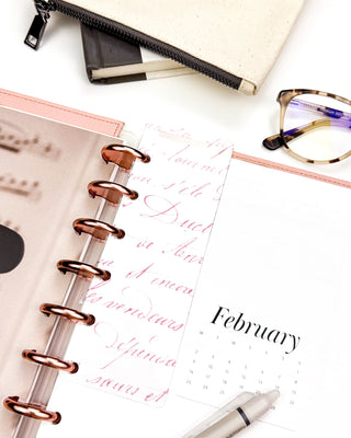 Romantic pink colored French handwriting on a glass clear plastic Page Finder. On a discbound planner with Rose Gold discs and Blush Snap-on Vegan Leather Cover on a desk flat lay. 