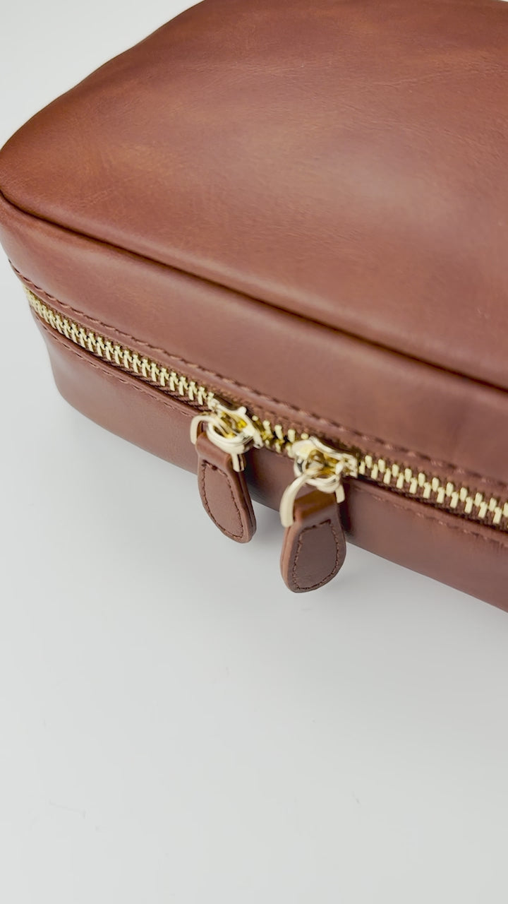 Close-up video of a warm brown leather zippered planner case featuring smooth leather with natural grain and gold-tone dual zipper hardware. The camera slowly glides across the corner of the case, highlighting the rich texture, precise stitching, and polished zipper pulls against a clean white background.