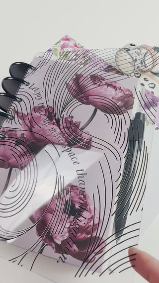 Close-up of a clear planner cover with black abstract line artwork layered over a mauve floral planner cover. The planner is styled with black discs, coordinating floral inserts, black-and-white washi tape, and a black pen.
