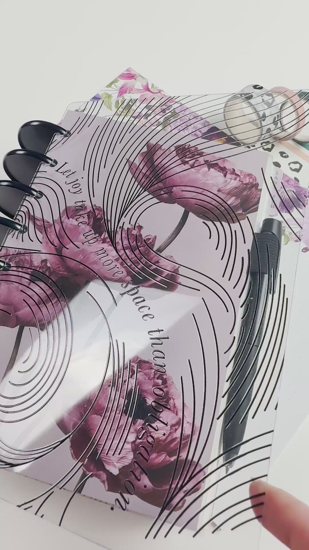 Close-up of a clear planner cover with black abstract line artwork layered over a mauve floral planner cover. The planner is styled with black discs, coordinating floral inserts, black-and-white washi tape, and a black pen.